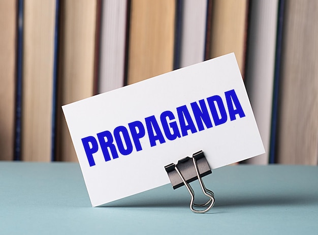 Белая карточка с текстом PROPAGANDA стоит на скрепке для бумаг на столе на фоне книг Defocus