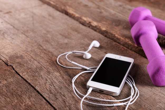 White cellphone with headphones and pink dumb bells on wooden background