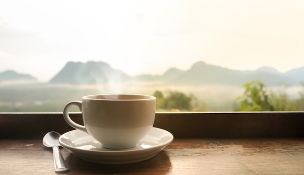 ぼやけた山の風景の上の日光と朝の木製のテーブルに白いセラミックコーヒーカップ