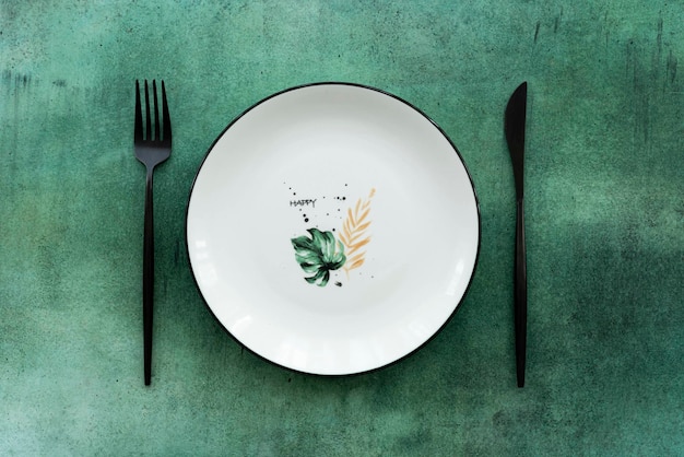 Белая керамическая тарелка с рисунком и столовые приборы на зеленом столе