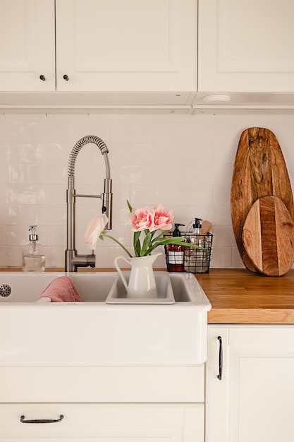 White ceramic sink with faucet hose and pink ranunculus flowers\
in it in a modern scandinavian style apartment. cozy home\
decoration concept. wooden counter with various kitchenware.