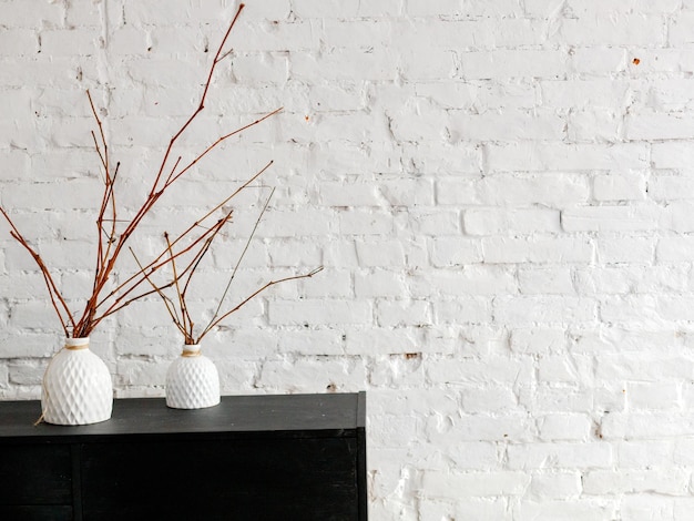 A white ceramic vase with dry branches on a black pedestal against a white brick wall Interior