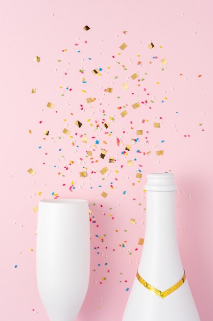 White champagne bottle and champagne glass with confetti on pink surface.