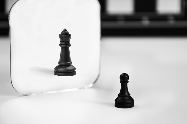 White chess pawn stands in front of the mirror, seeing reflection of queen. concept self-confidence