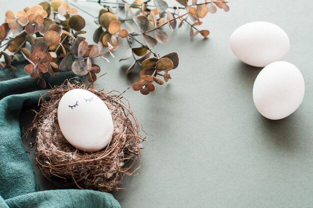 Белое куриное яйцо с глазами в гнезде, льняная салфетка, другие яйца и ветка эвкалипта на зеленом фоне, экологически чистая счастливая пасха
