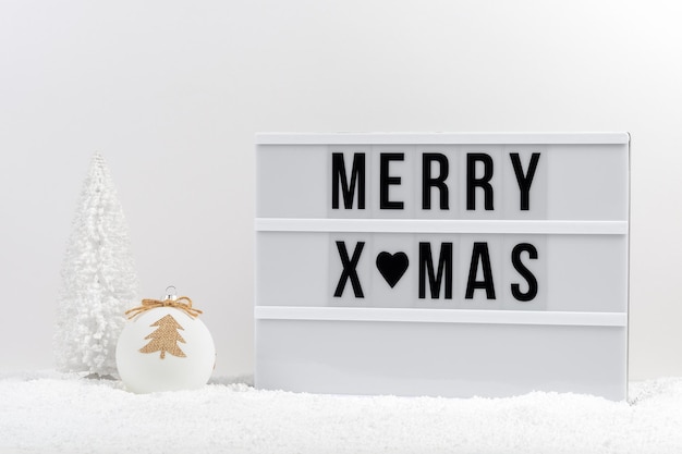 White Christmas tree Christmas bauble and lightbox with text MERRY XMAS on a table with snow