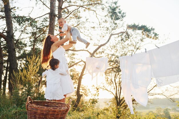 White clothes hanging on the rope to dry Young mother with her little daughter and son is outdoors in the forest Beautiful sunshine