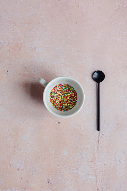 White coffee cup filled with candy sprinkles and a black spoon on a pink aged concrete background.