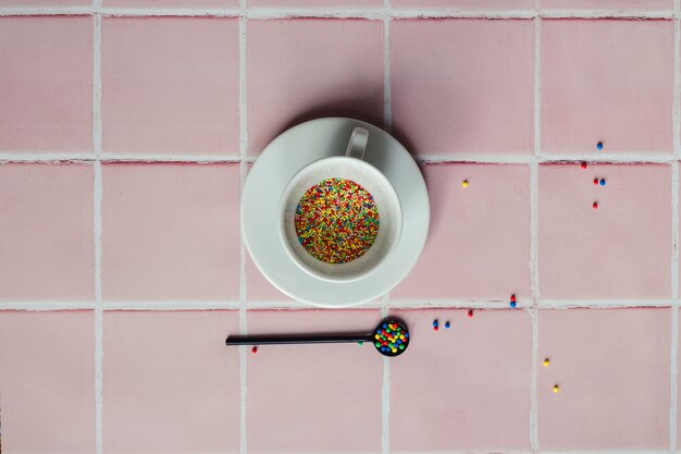 White coffee cup filled with candy sprinkles and a black spoon on a pink tile counter background