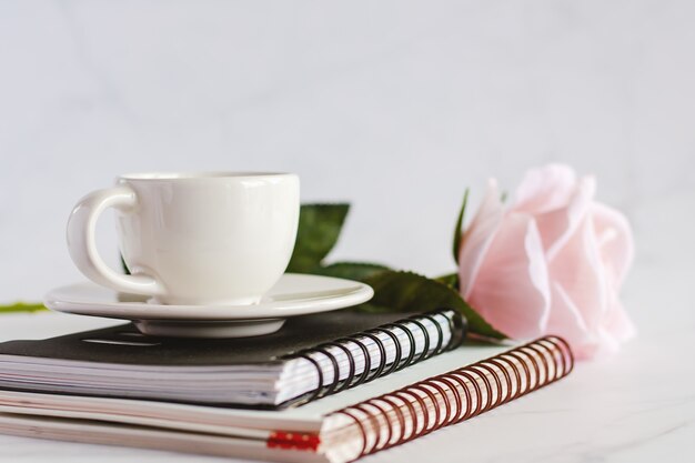 White coffee cup on spiral notebooks with sweet pink rose against white marble background