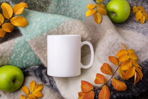 Mockup di tazza da caffè bianco con coperta di lana, mele verdi e foglie gialle autunnali. tazza vuota mock up per la promozione del design, modello in stile