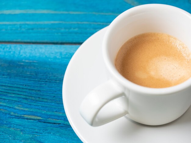 A white coffee mug with a full espresso is set on a saucer and on a blue wooden background. Coffee, energy drink. Delicious foam