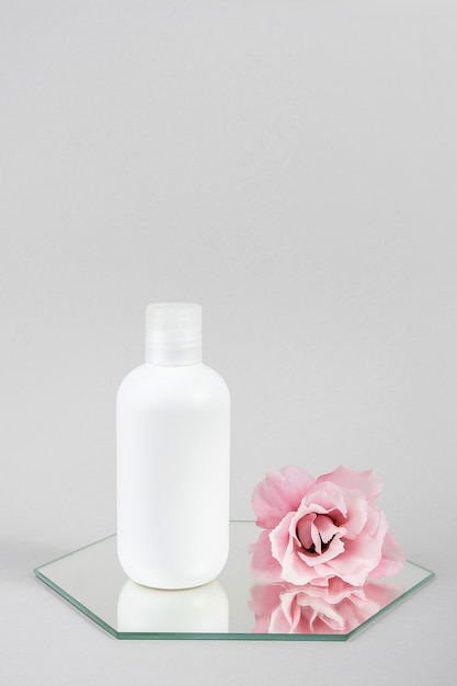 White cosmetic blank bottle and pink flower on mirror, gray background. Natural Organic Spa Cosmetic Beauty Concept Mockup, Front view.