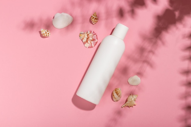 White cosmetic bottle with face cream or lotion and telana against a pink background with seashells and shadows Sun cream summer cosmetics