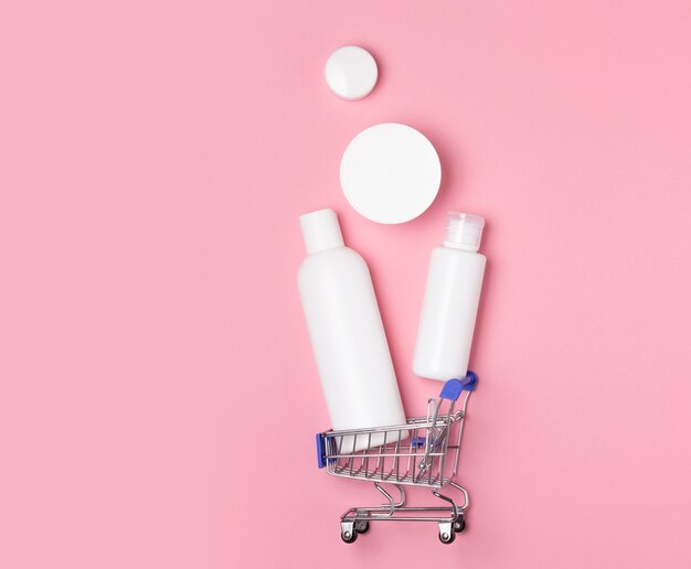 Белые косметические баночки лежат в корзине для покупок на розовом фоне. Интернет-магазины для дома. Покупка средств дезинфекции, стирки и ухода.