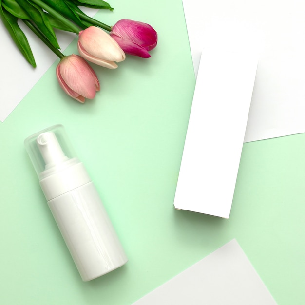 White cosmetic products and pink tulip flower on mint green and white paper background. 