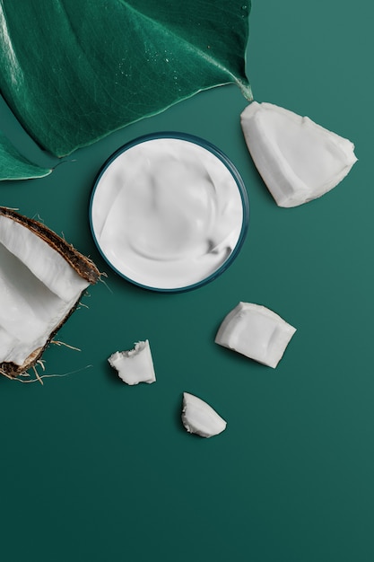 White cream in a jar on an exotic tropical leaf with coconut on trendy dark green Eden. beauty and health care concept. Minimalistic flat lay with copy space. Top view