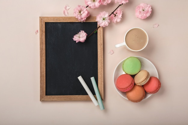 Белая чашка капучино с цветами сакуры, макароны, меловой доске на пастельном розовом фоне