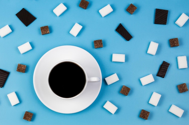 White cup of coffee brown and white sugar cubes and chocolate prieces on blue background