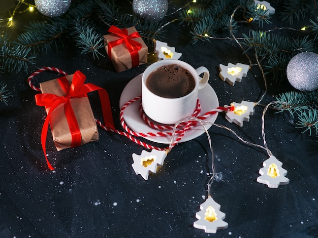 夕方の暗いテーブルに白い一杯のコーヒー、クリスマスプレゼント、クリスマスツリーの形をした花輪。家庭的な休日の雰囲気