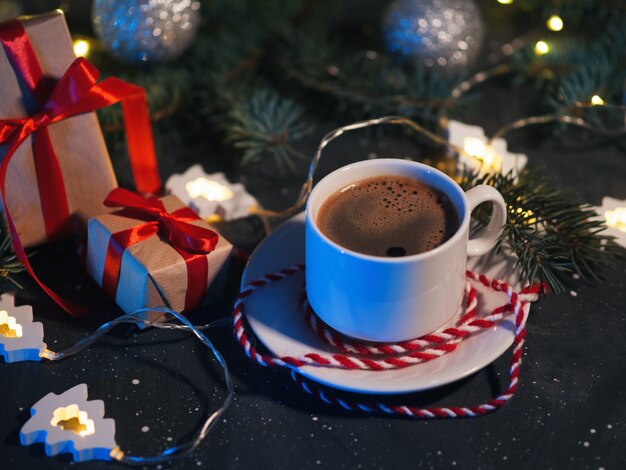 夕方の暗いテーブルに白い一杯のコーヒー、クリスマスプレゼント、クリスマスツリーの形をした花輪。家庭的な休日の雰囲気