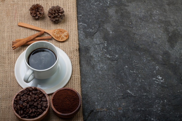 White cup coffee, coffee beans in wood cup , pine on burlap on black stone  textured