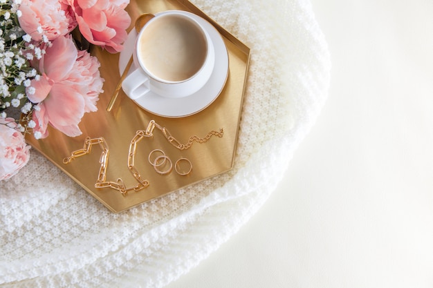 White cup of coffee and decorations on a golden tray in the nordic style. Pink peonies. A white plaid lies on a leather sofa.