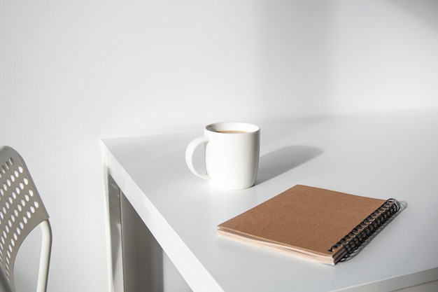 White cup of coffee a notebook on a white table there is a chair next to it