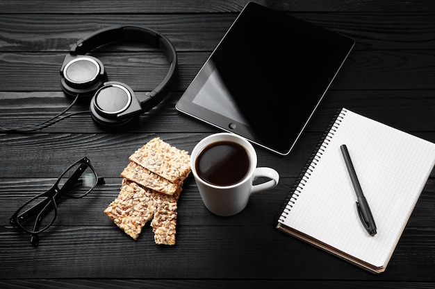 White cup of coffee notes book headphones and tablet on office wooden black working desktop