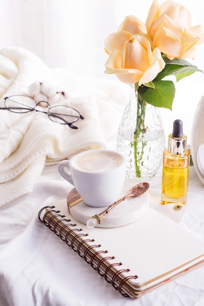 White cup of coffee and roses with notebook on white bed and plaid, cozy morning light. 