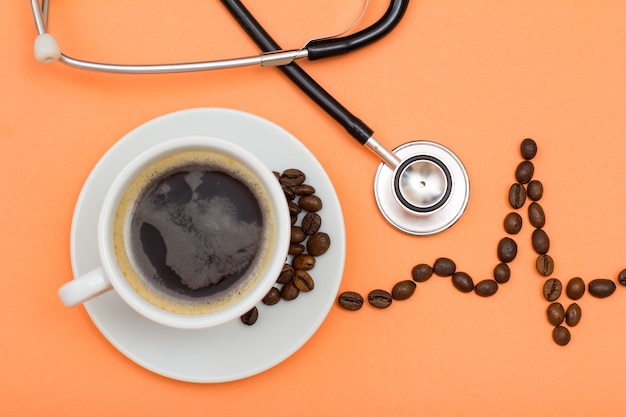 White cup of coffee on saucer with coffee beans folded in the form of a cardiogram and phonendoscope on peach colored background. Top view