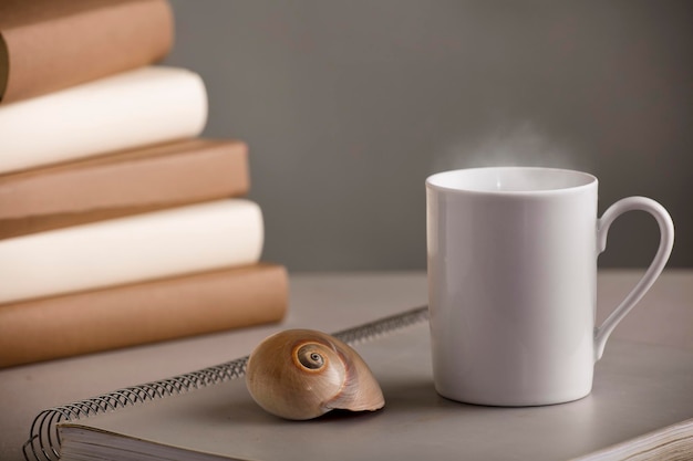 white cup of coffee with hot steam on the desk with nautilus shell and books grey background