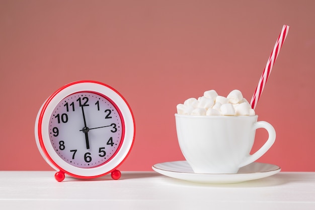 Una tazza bianca piena di marshmallow e una sveglia rossa su un tavolo bianco su sfondo rosa. il concetto di una bella mattina.