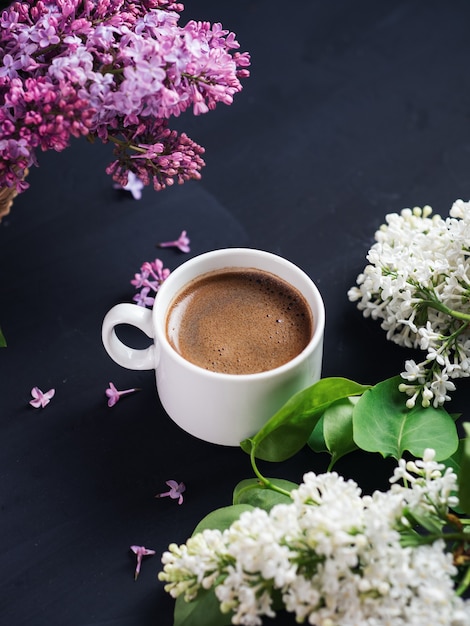 A white cup of fragrant coffee on a dark stone table with branches of purple and white lilacs. Spring morning and mood