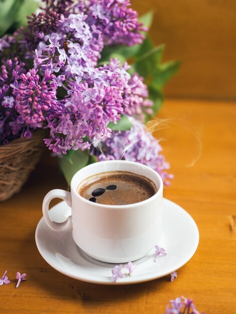 A white cup of fragrant coffee on a wooden table, a blooming lilac in a basket. May has come