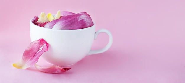 White cup full of fresh flowers petals of a tulips on a pink background banner