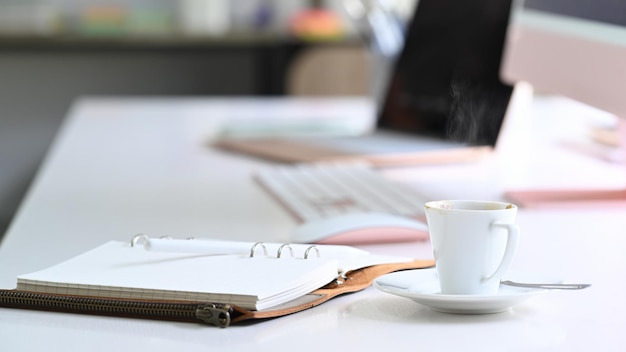 White cup of hot beverage and notebook on white office desk