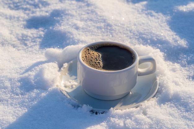 White cup of hot coffee on a bed of snow and white background, close up. Concept of christmas winter morning