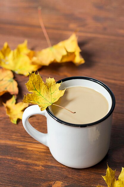 White cup of hot coffee with autumn leaves on wood background