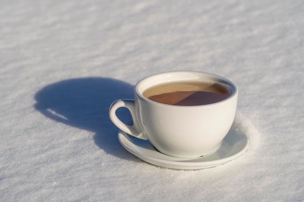 White cup of hot tea on a bed of snow and white background close up