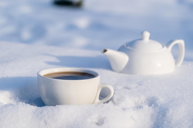 White cup of hot tea and teapot on a bed of snow and white background close up