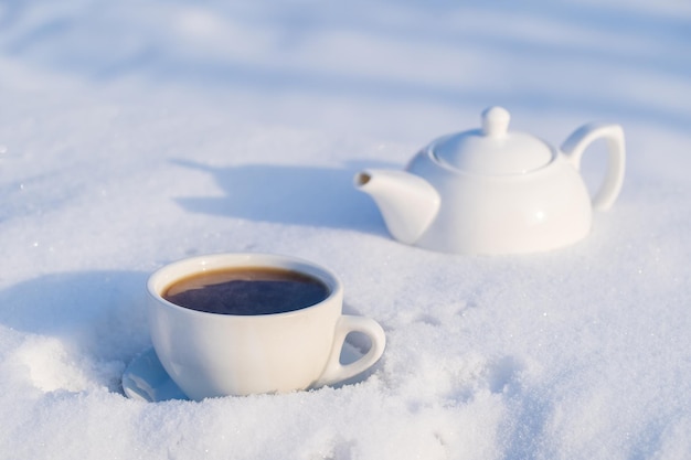 White cup of hot tea and teapot on a bed of snow and white background close up