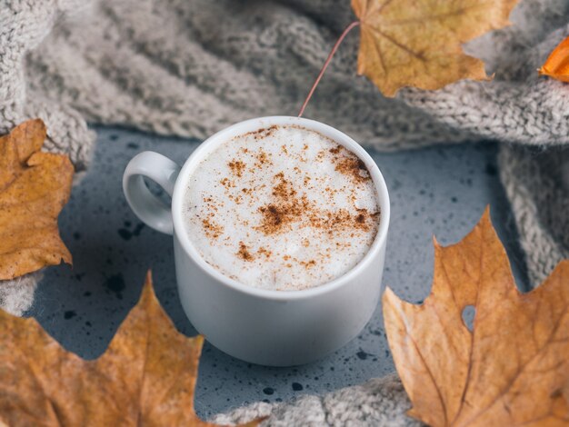 White Cup of latte coffee on gray table with a knitted scarf and a dry autumn maple leaves. Home atmosphere and comfort in cold weather