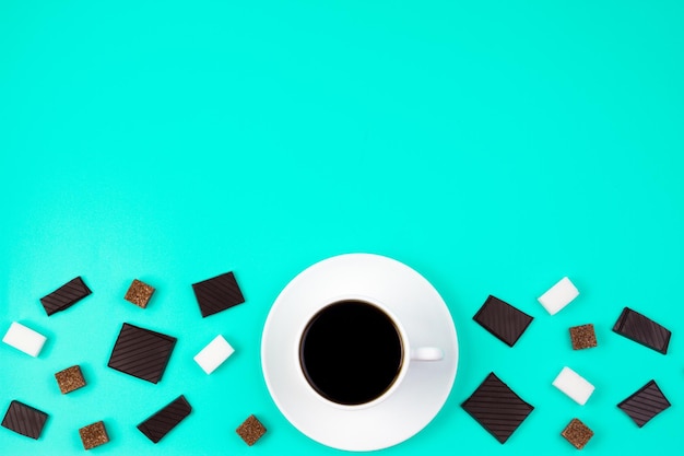 Фото Белая чашка кофе, коричневые и белые кубики сахара и кусочки шоколада на бирюзовом фоне