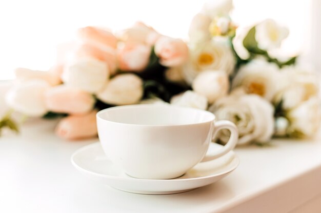 A white cup on a saucer stands against a background of white tulips