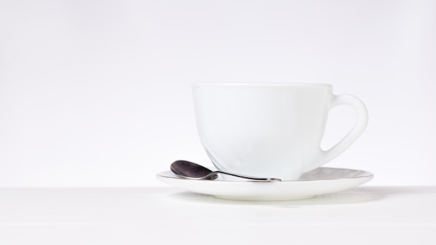 Photo a white cup for tea or coffee and a metal spoon on a white table on a gray background. dishes for tea and coffee.