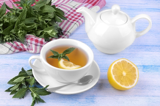 White cup of tea with mint and lemon and a kettle on a blue wooden background