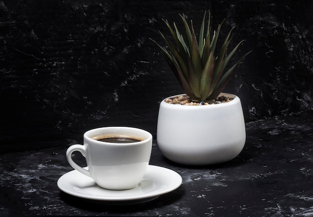 White cup with black aromatic coffee on a black abstract background with a potted flower in the background.