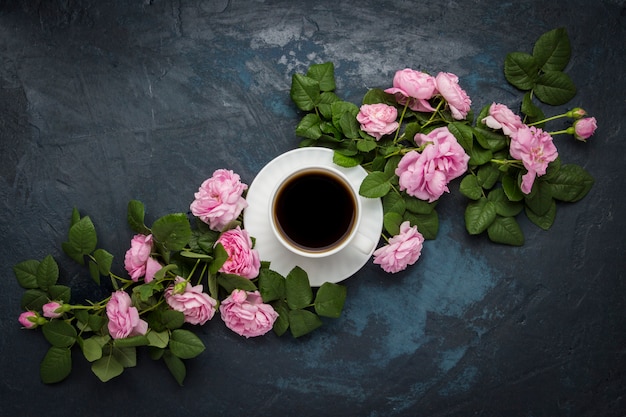 White cup with black coffee and pink roses on a dark blue surface. Flat lay, top view