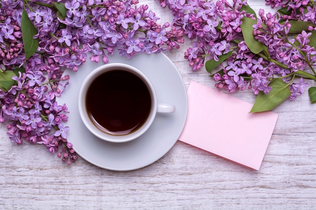 White cup with coffee or tea, note with wishes, a bouquet of lilacs on a wooden background. Violet spring flowers. Morning composition.